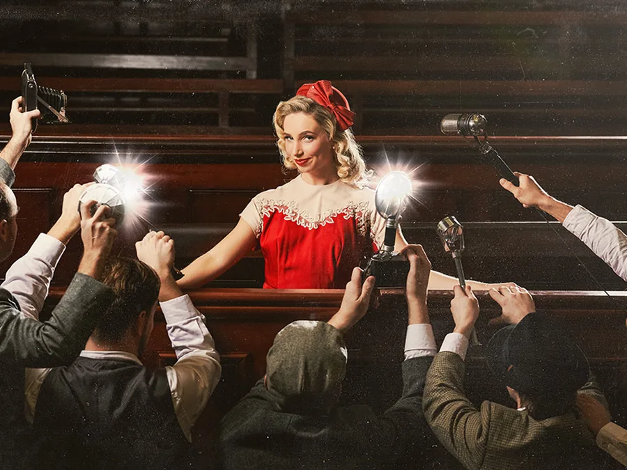A woman in a red dress in a courtroom dock, surrounded by press photographers with flashbulbs